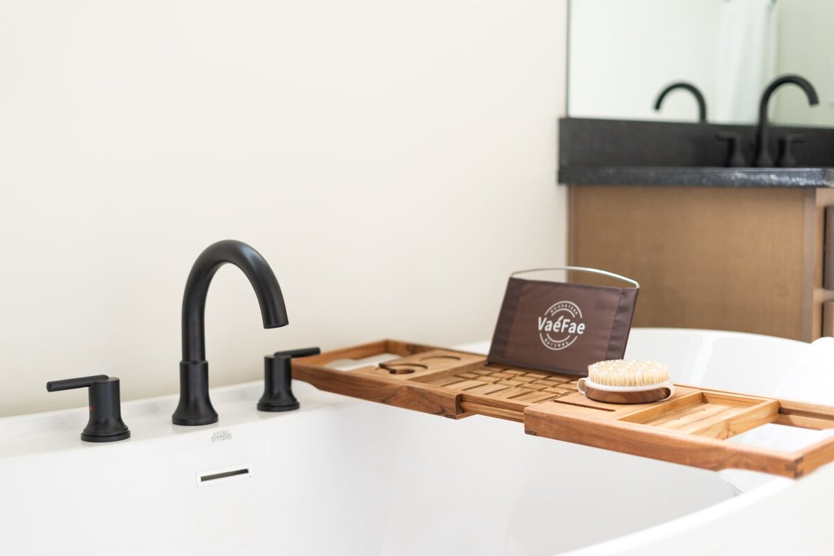 A detailed close-up of a wooden bath caddy resting on the edge of a white soaking tub, featuring compartments for a book, glass, and a body brush. Black modern bath fixtures are visible on the tub's rim.