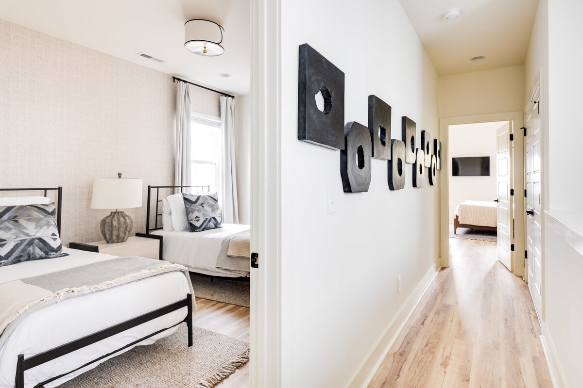 A view from a hallway into a bright bedroom with two twin beds, white bedding, and gray patterned pillows, featuring a light patterned accent wall and light wood floors. The hallway displays a series of abstract black wall art pieces.