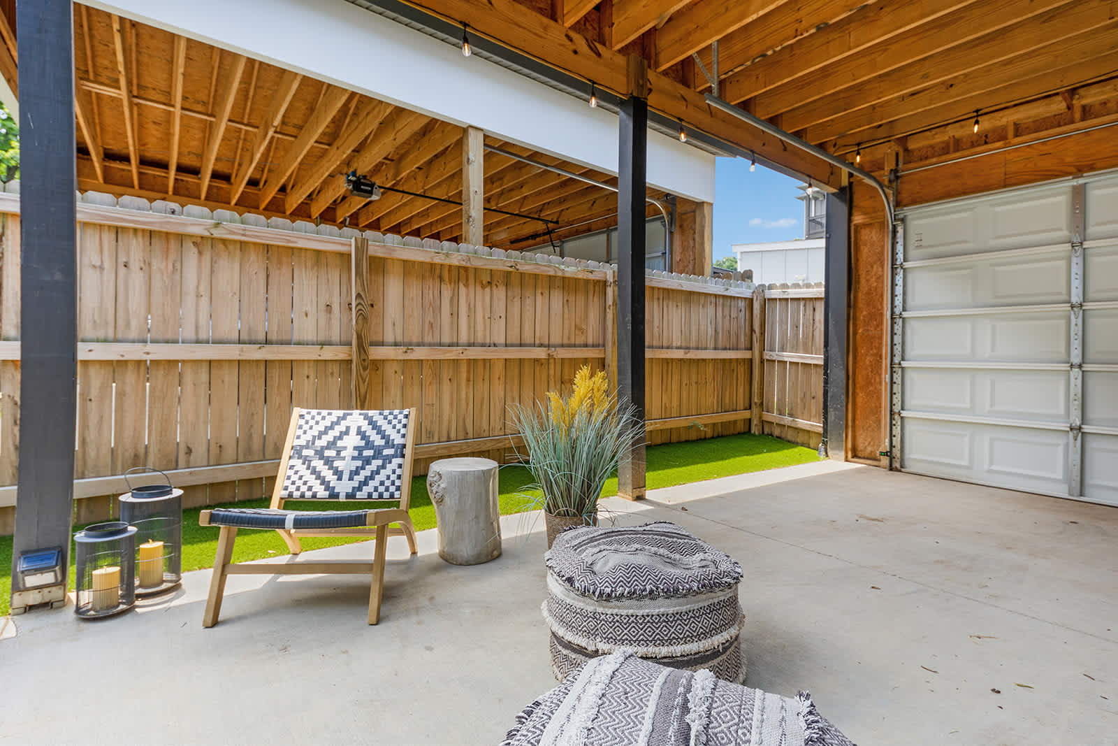 A spacious covered patio area with concrete flooring features a black and white patterned chair, a wooden stump side table, and a large patterned pouf, set against a wooden fence and artificial turf. A white garage door is visible in the background.