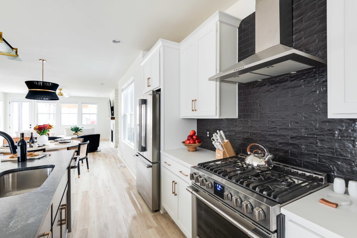Kitchen view showing white cabinets, a stainless steel range with a matching hood, a textured black subway tile backsplash, and a dark kitchen island with a sink, illuminated by recessed lighting.