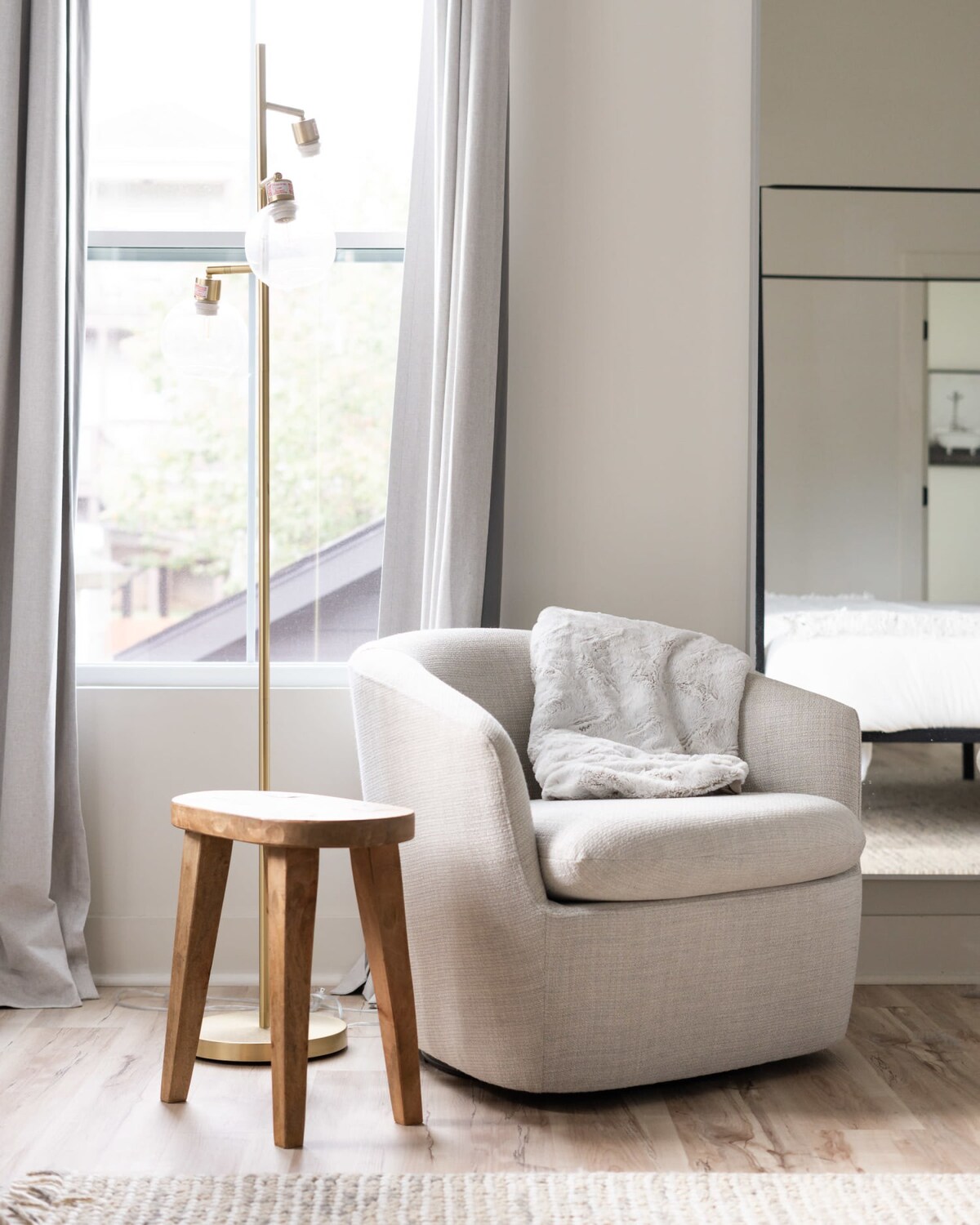 A cozy sitting area in a bedroom, featuring a light gray upholstered armchair with a textured throw blanket, next to a rustic wooden stool. A modern brass floor lamp with globe lights stands by a window with light gray curtains.