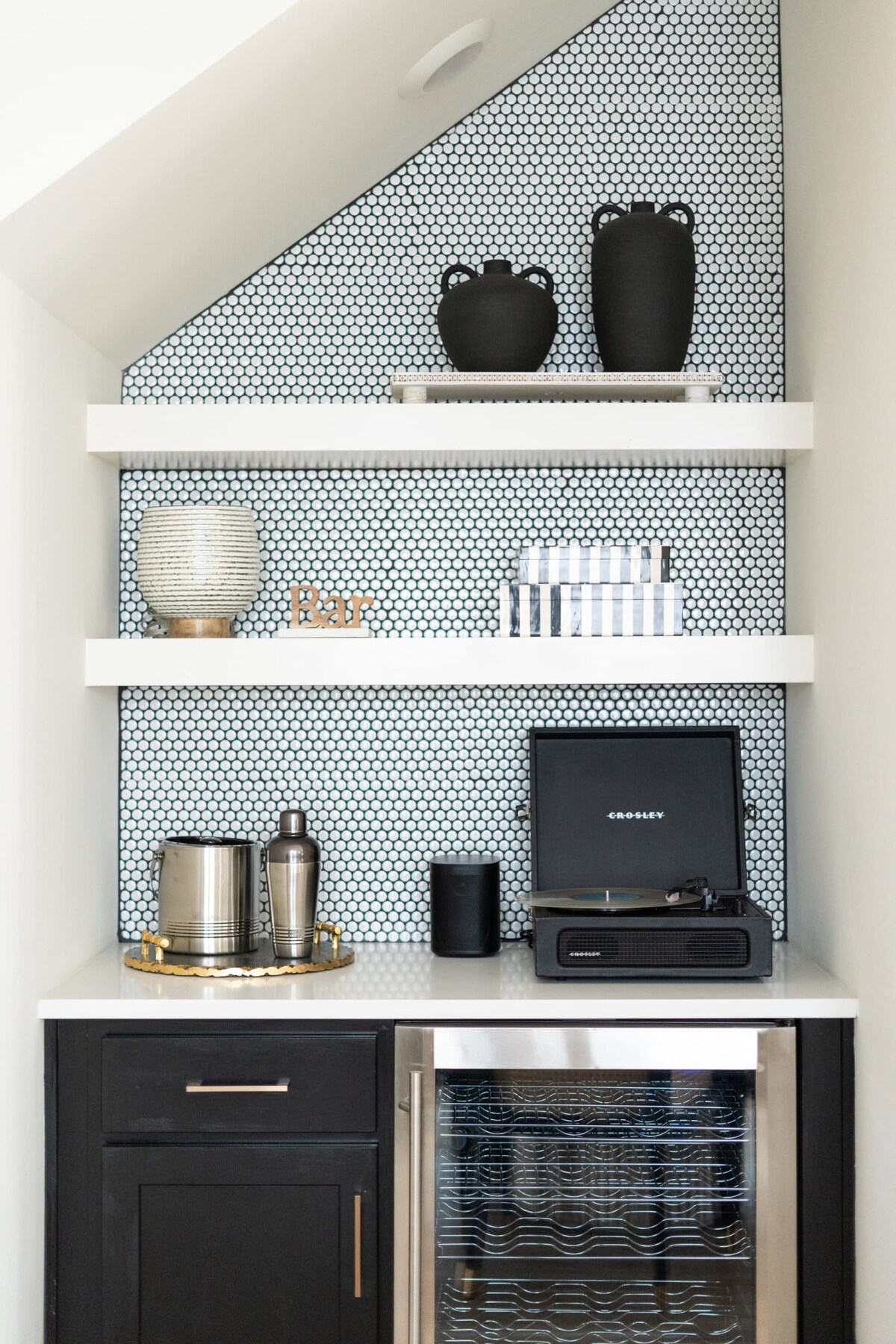 A stylish wet bar nook with white floating shelves against a black and white penny tile backsplash, featuring black decorative vases and a record player on the white countertop.