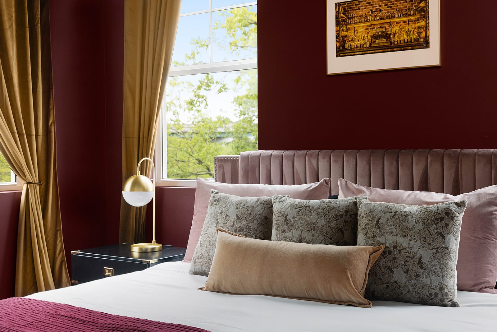 A close-up of the bed in a bedroom with vibrant red walls, showcasing a tufted headboard, an array of soft pink and brown pillows, and a modern gold bedside lamp on a sleek black nightstand.