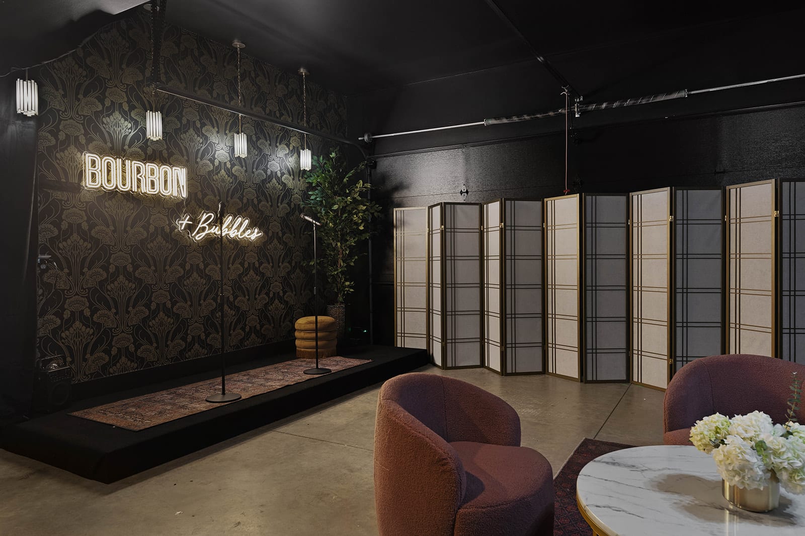 A dark, speakeasy-themed entertainment space features black and gold patterned wallpaper, a neon "Bourbon + Bubbles" sign, a small stage, and a decorative room divider.