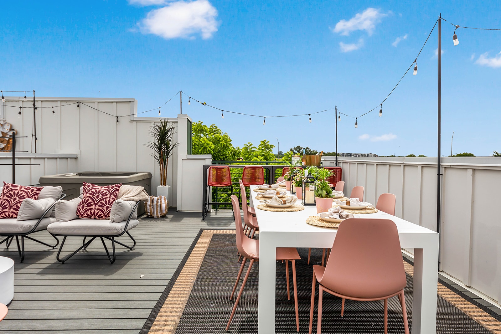 A spacious rooftop deck featuring a hot tub, comfortable outdoor seating with decorative pillows, and a dining area with a white table and pink chairs. String lights are strung across the deck, and the sky is bright blue.