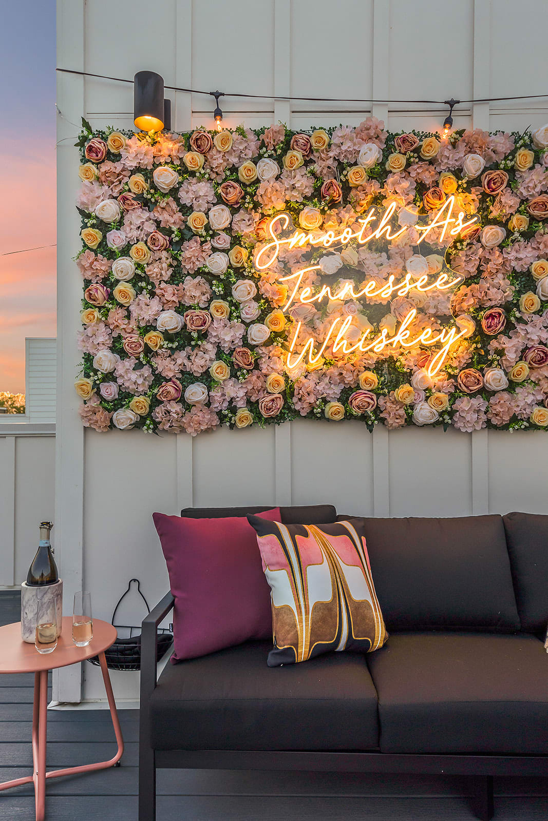 A close-up of the rooftop deck lounge area, showcasing a black outdoor sofa with colorful pillows, a pink side table with champagne, and a vibrant floral wall with a "Smooth As Tennessee Whiskey" neon sign.