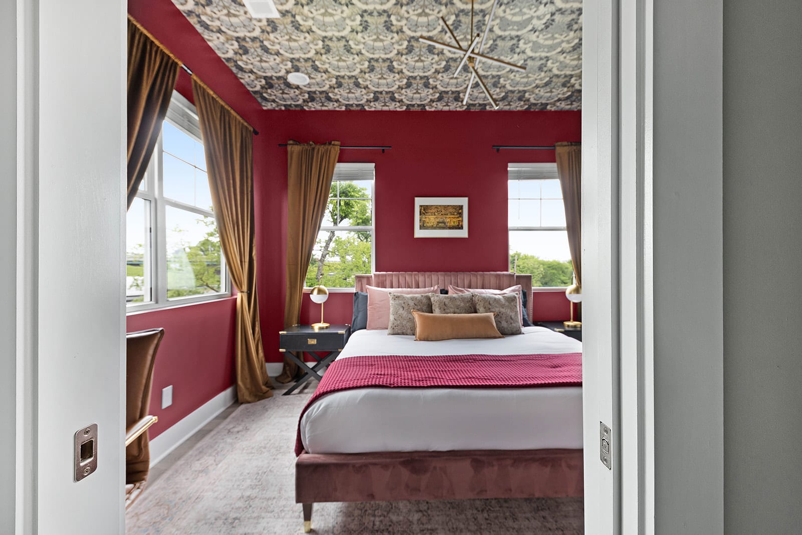 A luxurious bedroom with bold red walls and a unique patterned ceiling, featuring a bed with a plush tufted headboard and a deep red throw. Large windows are elegantly framed by rich gold curtains.