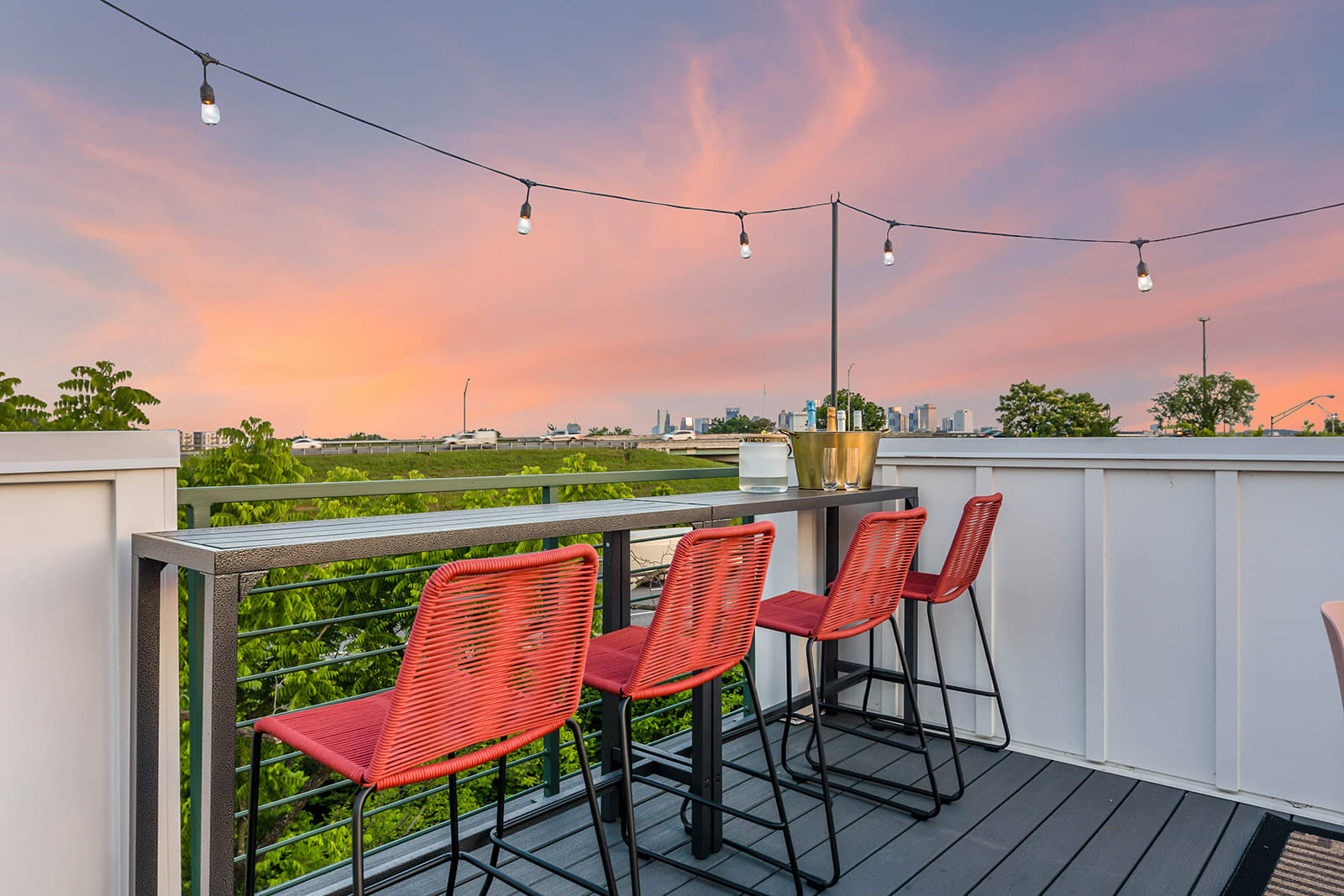 Rooftop deck featuring a long bar table with three red woven bar stools, string lights overhead, and a scenic sunset view of a distant city skyline and highway.