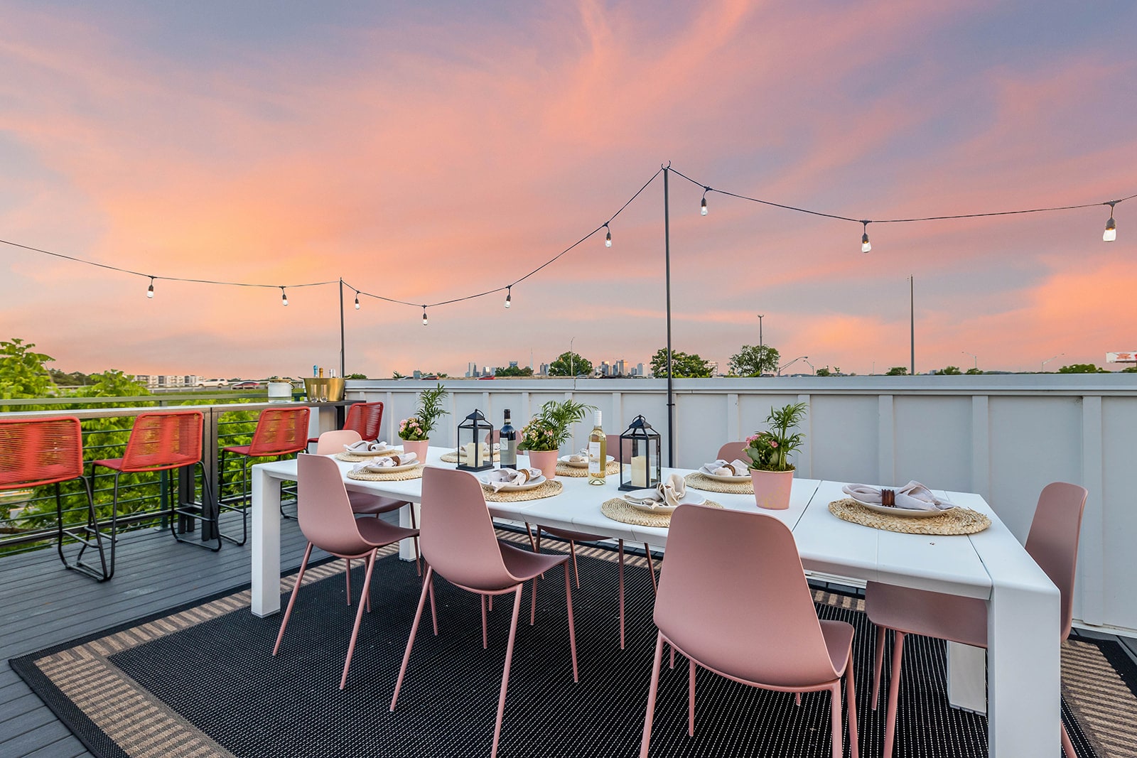 A spacious rooftop deck set for an evening meal, featuring a long white dining table with pink chairs, string lights overhead, and a stunning sunset sky with city views in the distance. The deck has dark grey flooring and white railings.
