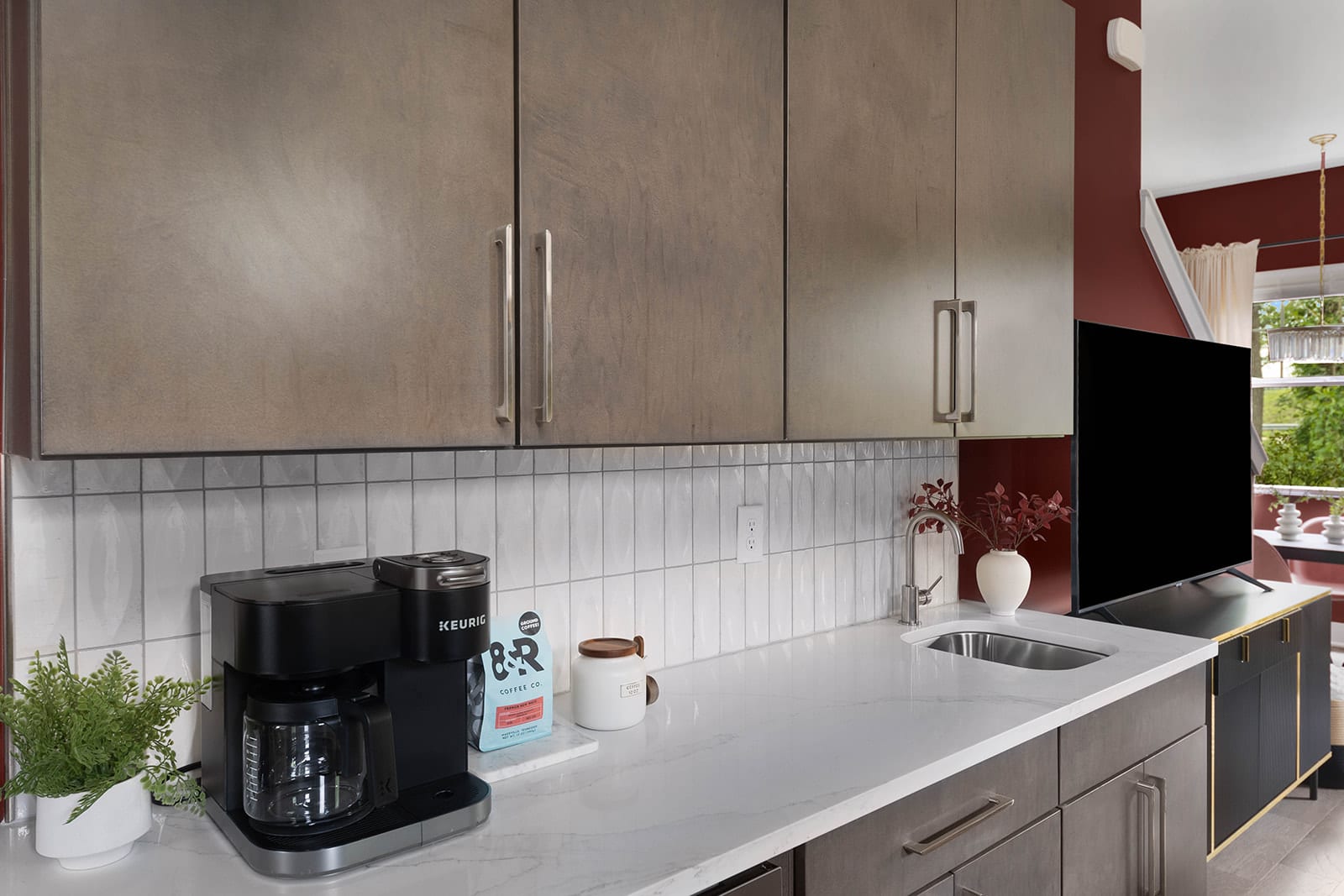 A close-up of a modern kitchen counter features light wood upper cabinets and a white subway tile backsplash. A black Keurig coffee maker, a coffee bag, and a white mug are neatly arranged on the pristine white quartz countertop.