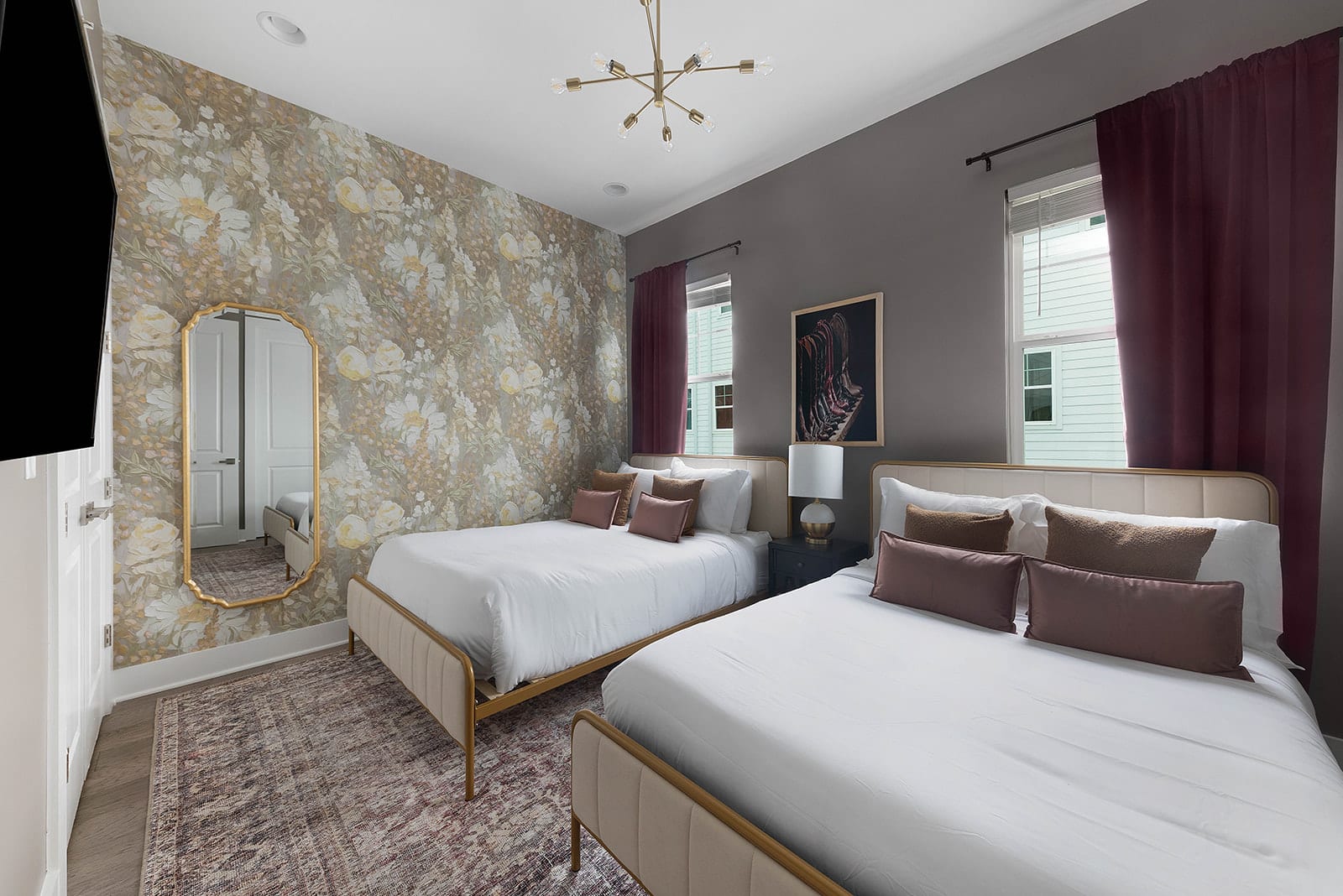 A stylish bedroom features two queen beds with cream upholstered headboards and gold frames, set against a floral wallpaper accent wall. The room includes a large gold-framed mirror, a modern chandelier, and burgundy curtains.