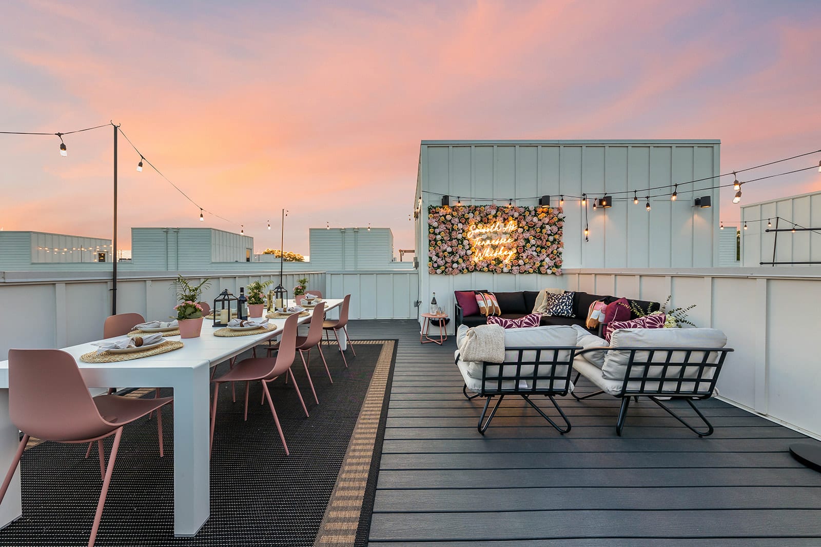 A spacious rooftop deck features both a white outdoor dining table with pink chairs and a lounge area with a black sectional sofa and fire pit, all under string lights with a floral neon sign and sunset views.