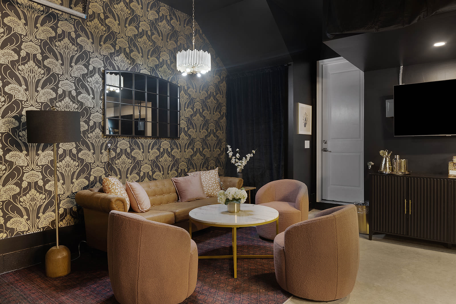 A cozy living area with dark and gold patterned wallpaper features a tufted brown leather sofa with pink pillows, two plush armchairs, a round marble coffee table, and a large black framed mirror.