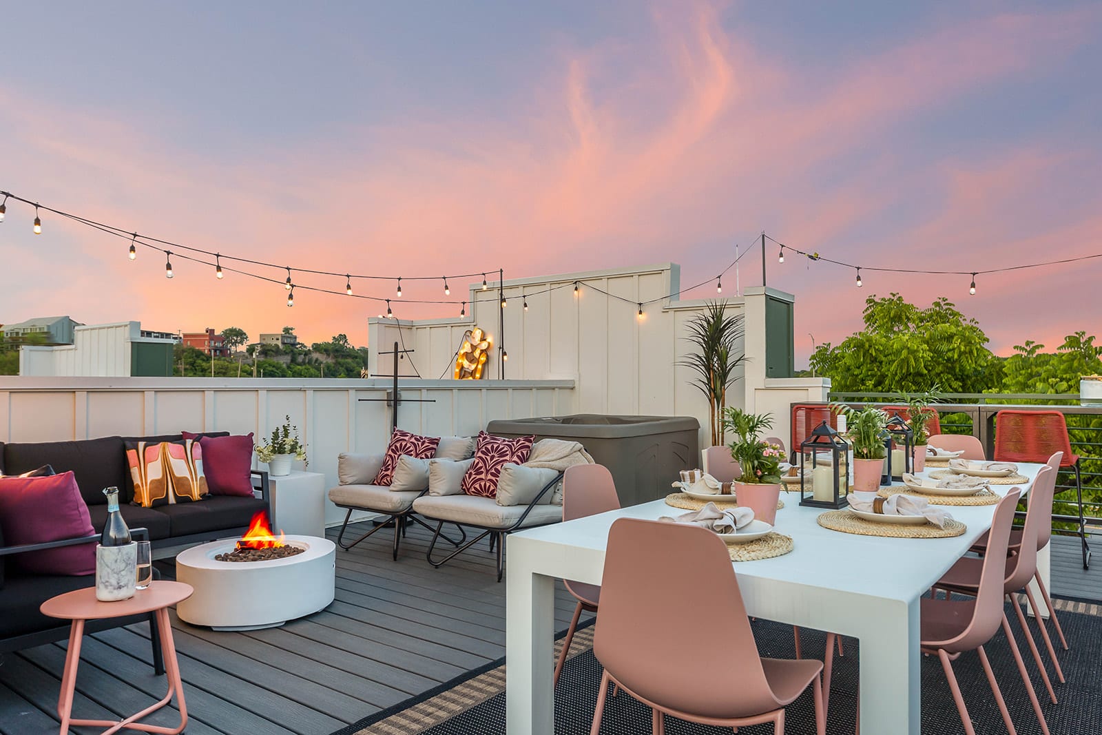 A vibrant rooftop deck at sunset features a long white dining table with pink chairs, a cozy seating area with a fire pit, and a hot tub, all illuminated by string lights, offering panoramic views.