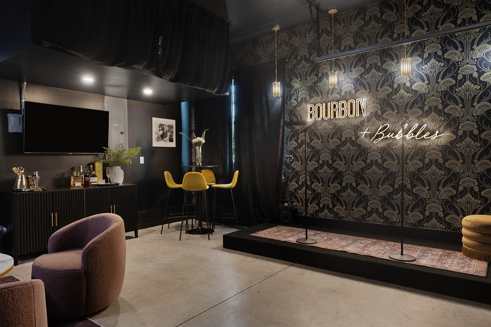 A dark, moody speakeasy-style room features black and gold patterned wallpaper, a neon "Bourbon + Bubbles" sign, a small stage with a rug, and a bar area with yellow bar stools and a TV.