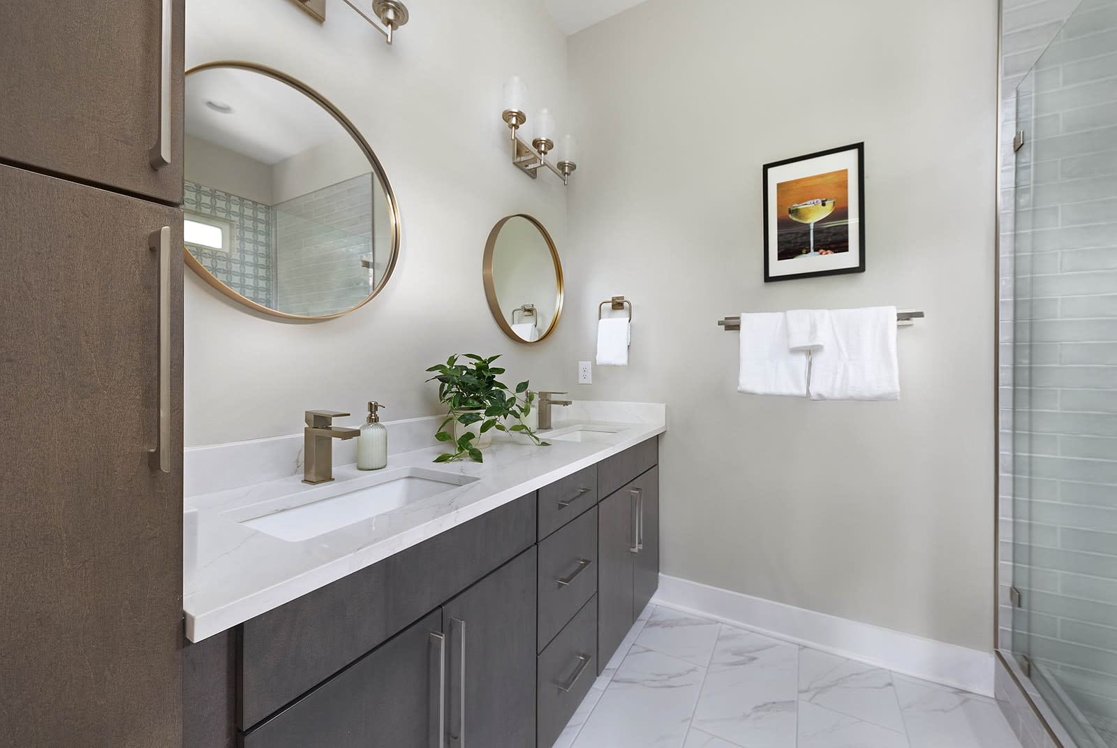 A modern bathroom with a double vanity featuring dark wood cabinets, white quartz countertops, and gold-toned faucets. Two round mirrors hang above the sinks, and a glass-enclosed shower with light gray subway tiles is visible.