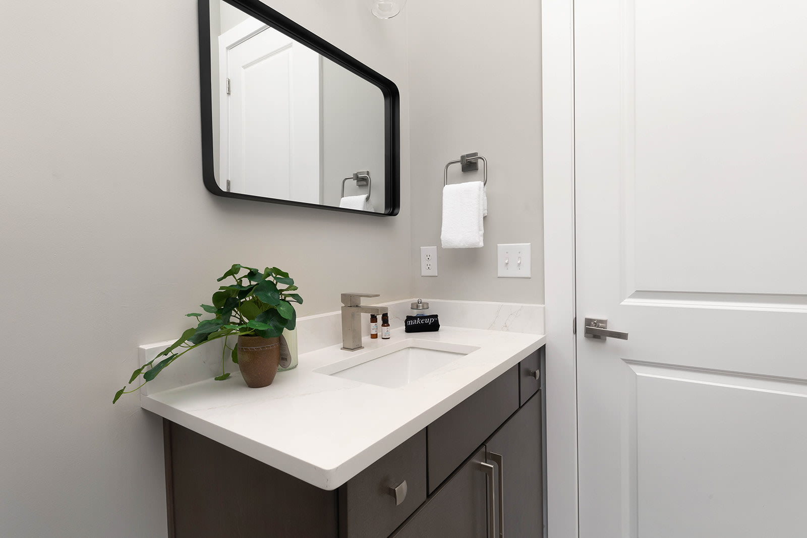 A bright bathroom with a single dark wood vanity topped with a white quartz counter and a rectangular black-framed mirror. A small green plant adds a touch of nature, and a white hand towel hangs on a silver rack.