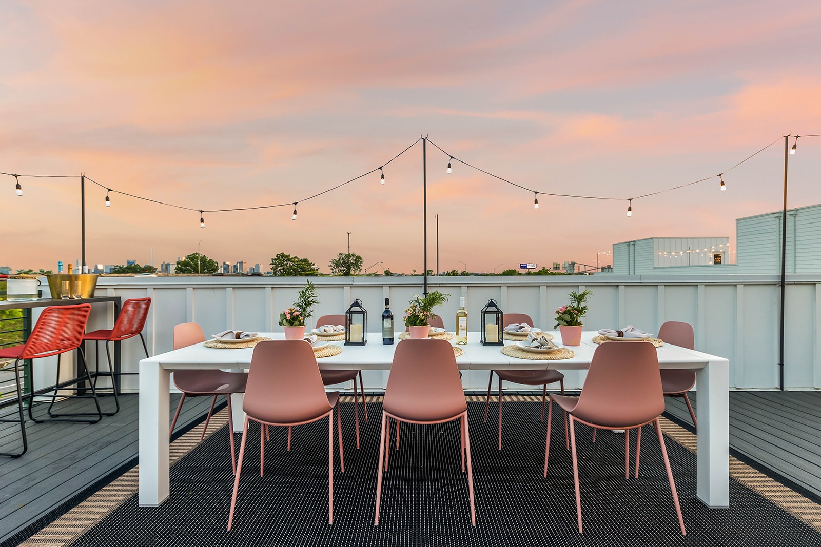 A white outdoor dining table with pink chairs is set on a dark rug on a rooftop deck, with string lights overhead and a vibrant sunset sky casting warm hues over the distant city skyline.