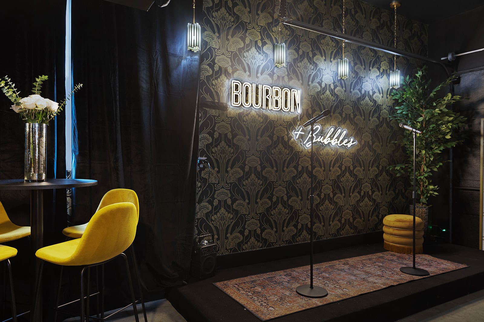 A close-up of the speakeasy's stage area showcases black and gold patterned wallpaper, a neon "Bourbon + Bubbles" sign, two microphones, and a small rug. Yellow bar stools are visible to the left.