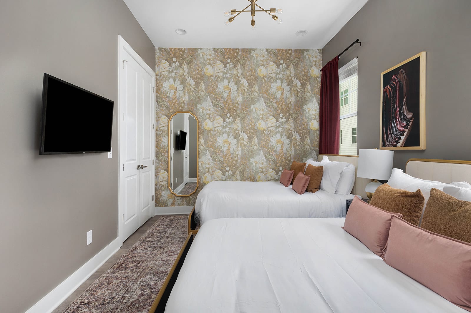 A stylish bedroom featuring two beds, one against a floral wallpaper accent wall and the other adjacent. The room includes a wall-mounted TV, a full-length mirror, and a patterned area rug on a light wood floor.