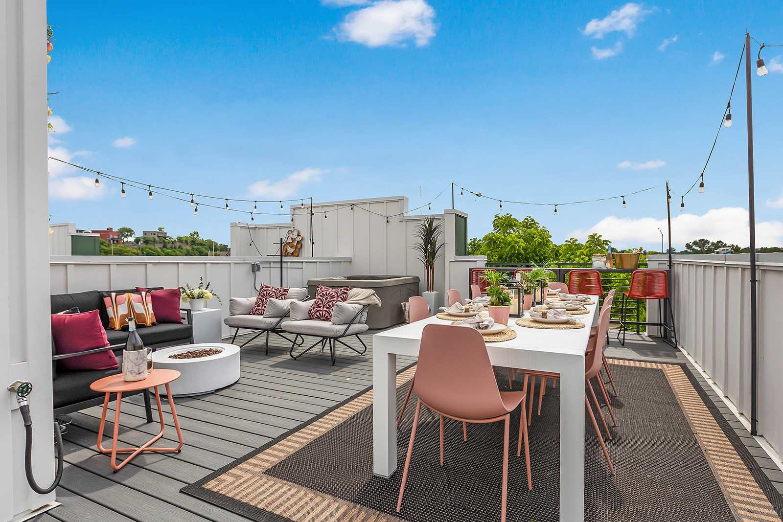 Expansive rooftop deck featuring a white dining table with pink chairs, a lounge area with a black sofa and armchairs, a circular fire pit, and string lights overhead, all under a bright blue sky.