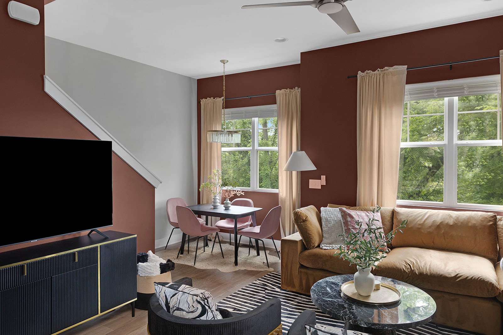 An open-concept living and dining area with a rich brown velvet sofa, a black marble coffee table, and a dining set with pink chairs. The space features a ceiling fan, large windows, and a black and gold TV console.