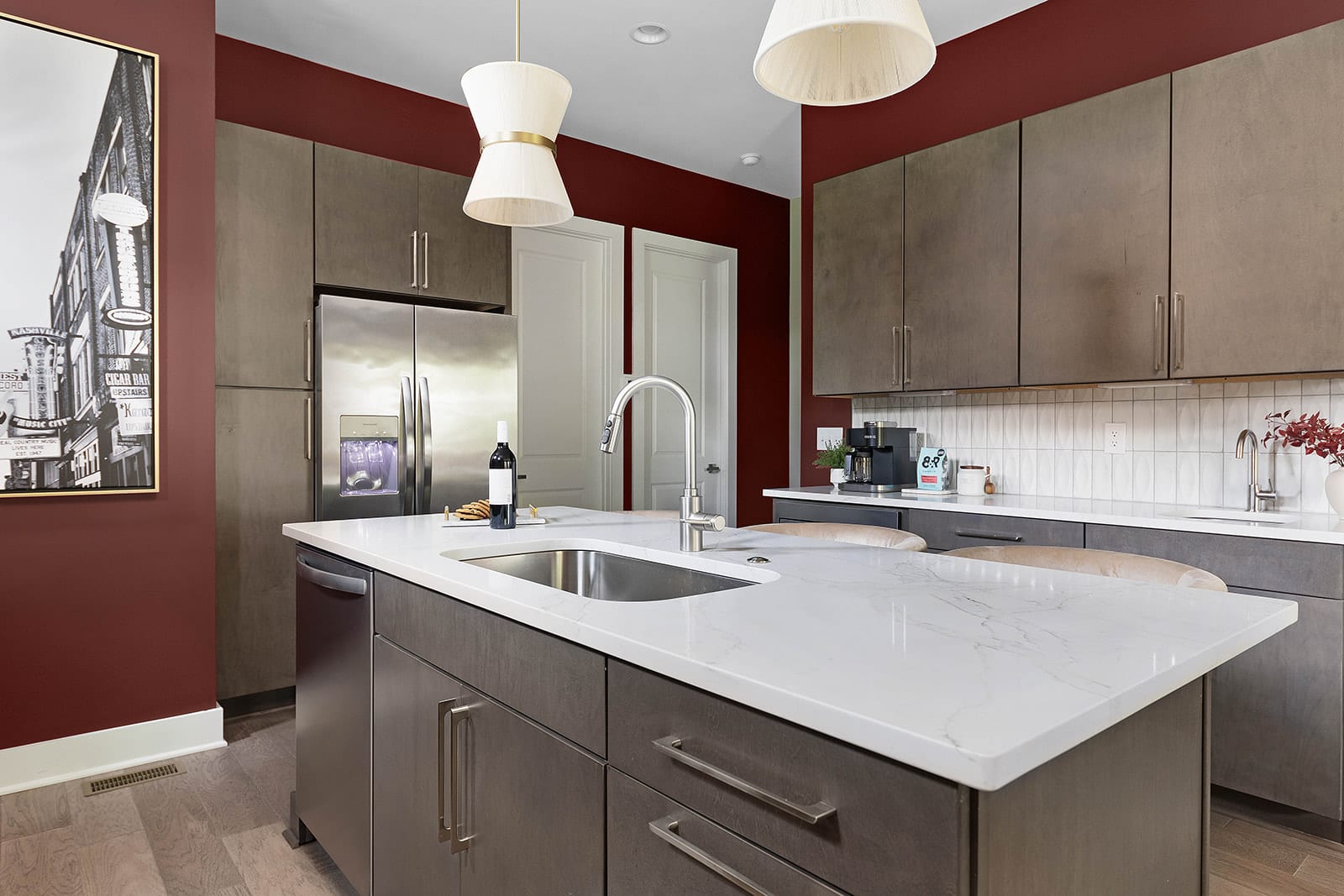 A spacious kitchen features a large island with a sink and modern faucet, dark gray cabinetry, and white quartz countertops. The room boasts a striking red accent wall, stainless steel appliances, and two contemporary pendant lights.