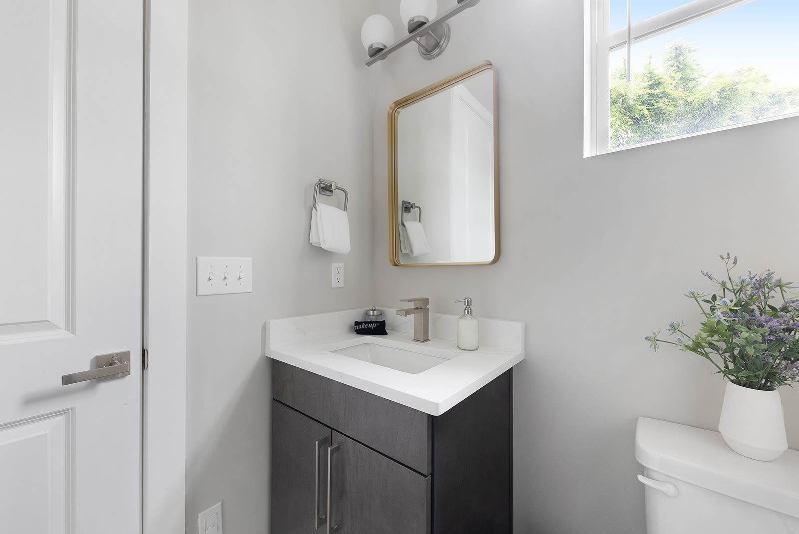 A modern powder room featuring a dark wood vanity with a white countertop and sink, a gold-framed rectangular mirror, and a window providing natural light. A white toilet is partially visible on the right.