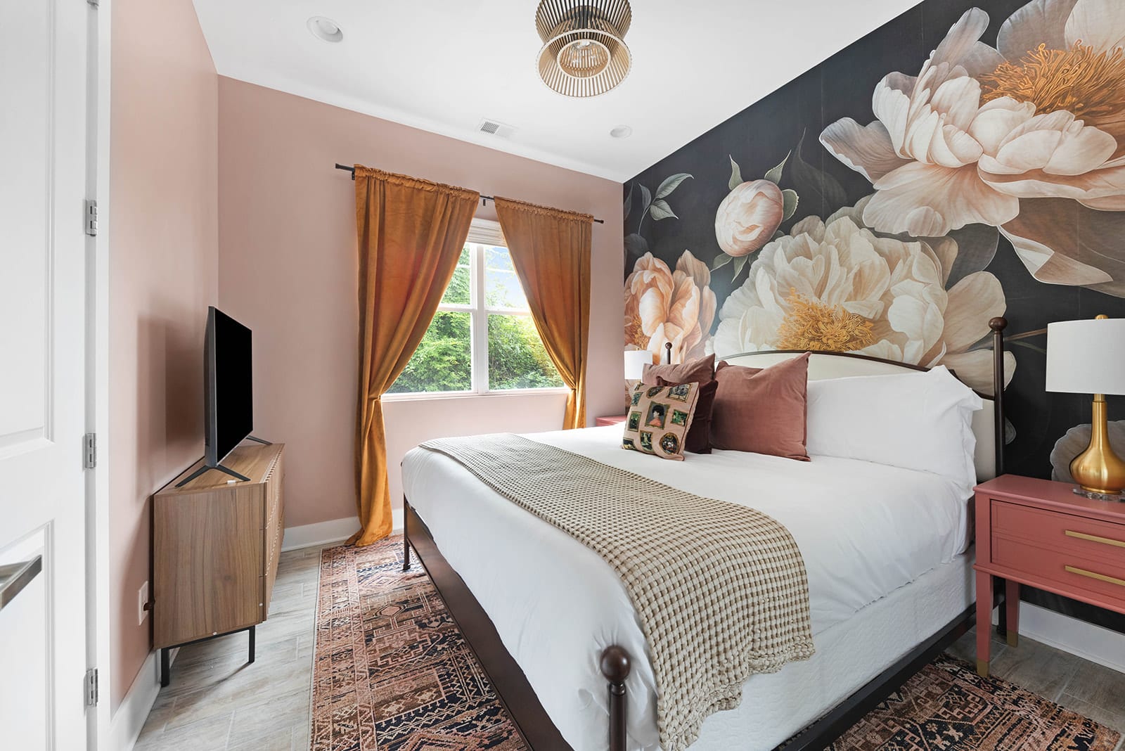 A cozy bedroom featuring a king-sized bed with a white duvet and a patterned throw, set against a dramatic dark floral accent wall. The room has light pink walls, a large window with orange curtains, and a wooden TV console.