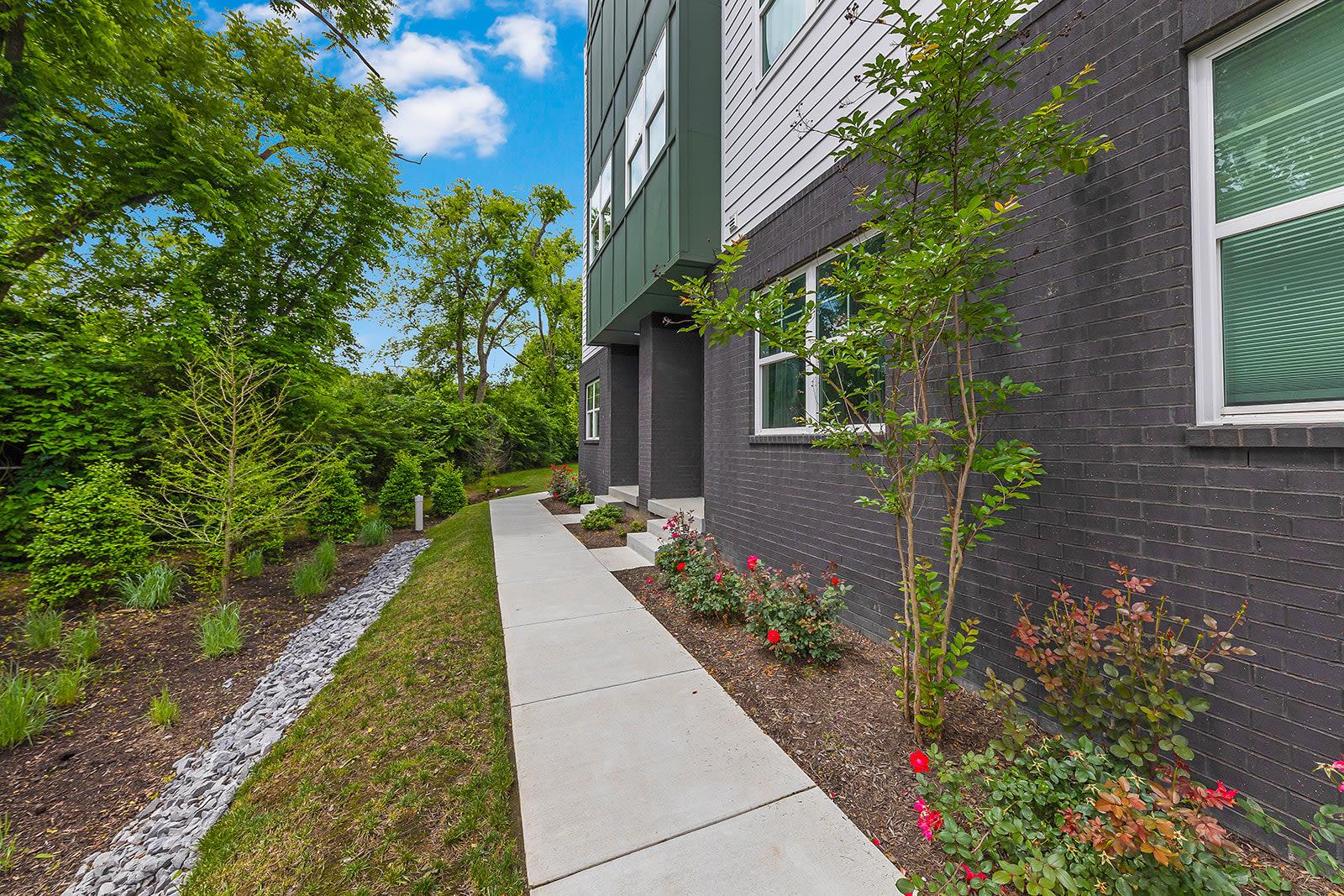 Exterior view of a modern townhouse with dark brick and green siding, a concrete walkway leading through landscaped garden beds with small trees and red flowers.