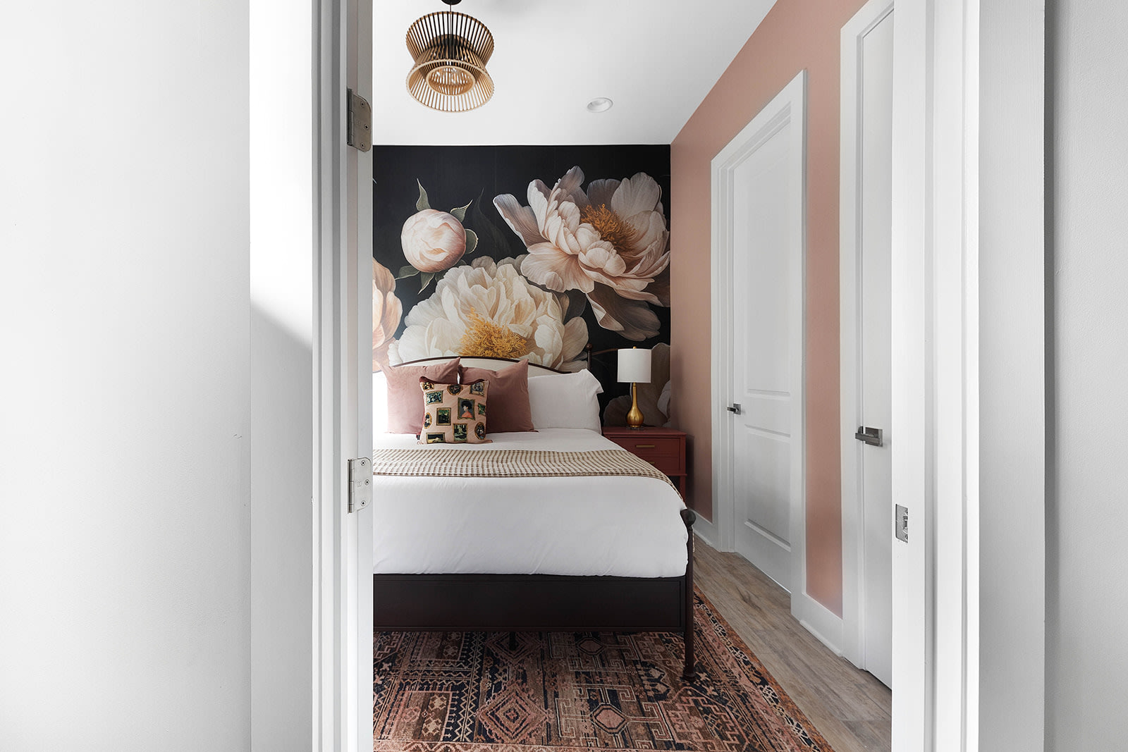 A charming bedroom featuring a striking large-scale floral mural on an accent wall behind the bed, complemented by an adjacent soft pink painted wall. A richly patterned rug covers the floor.