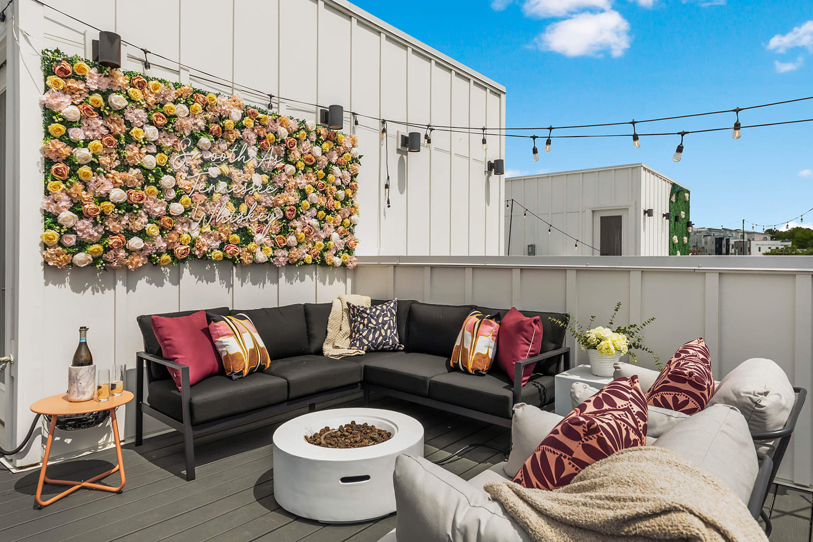 A stylish rooftop lounge area featuring a large black sectional sofa, a white fire pit, and additional seating with throw pillows. A vibrant floral wall art installation adds a pop of color, and string lights illuminate the space.