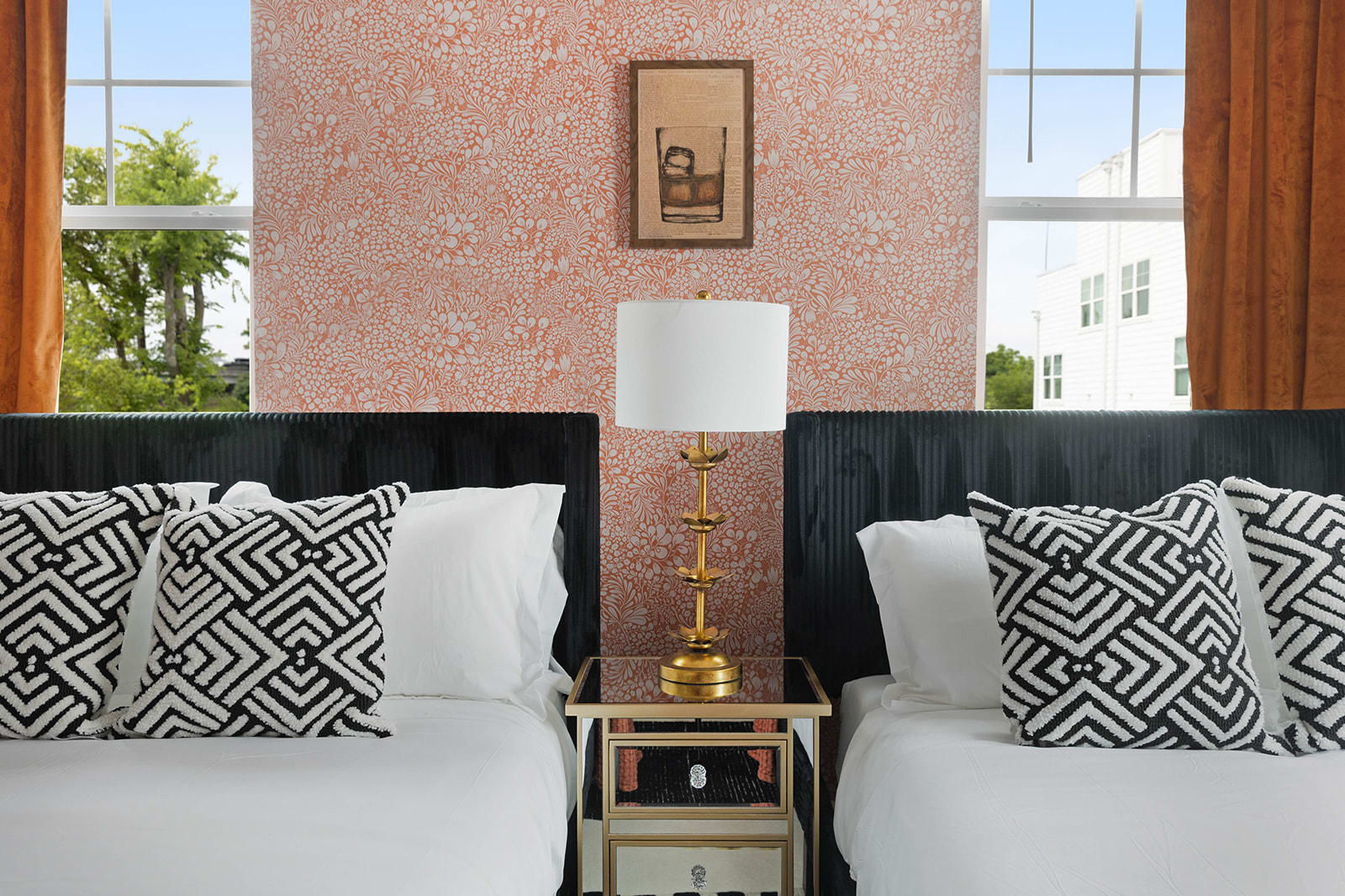 A close-up view between two beds in a bedroom, highlighting a chic gold lamp on a mirrored nightstand. The intricate patterned accent wall and a bright window are visible in the background.