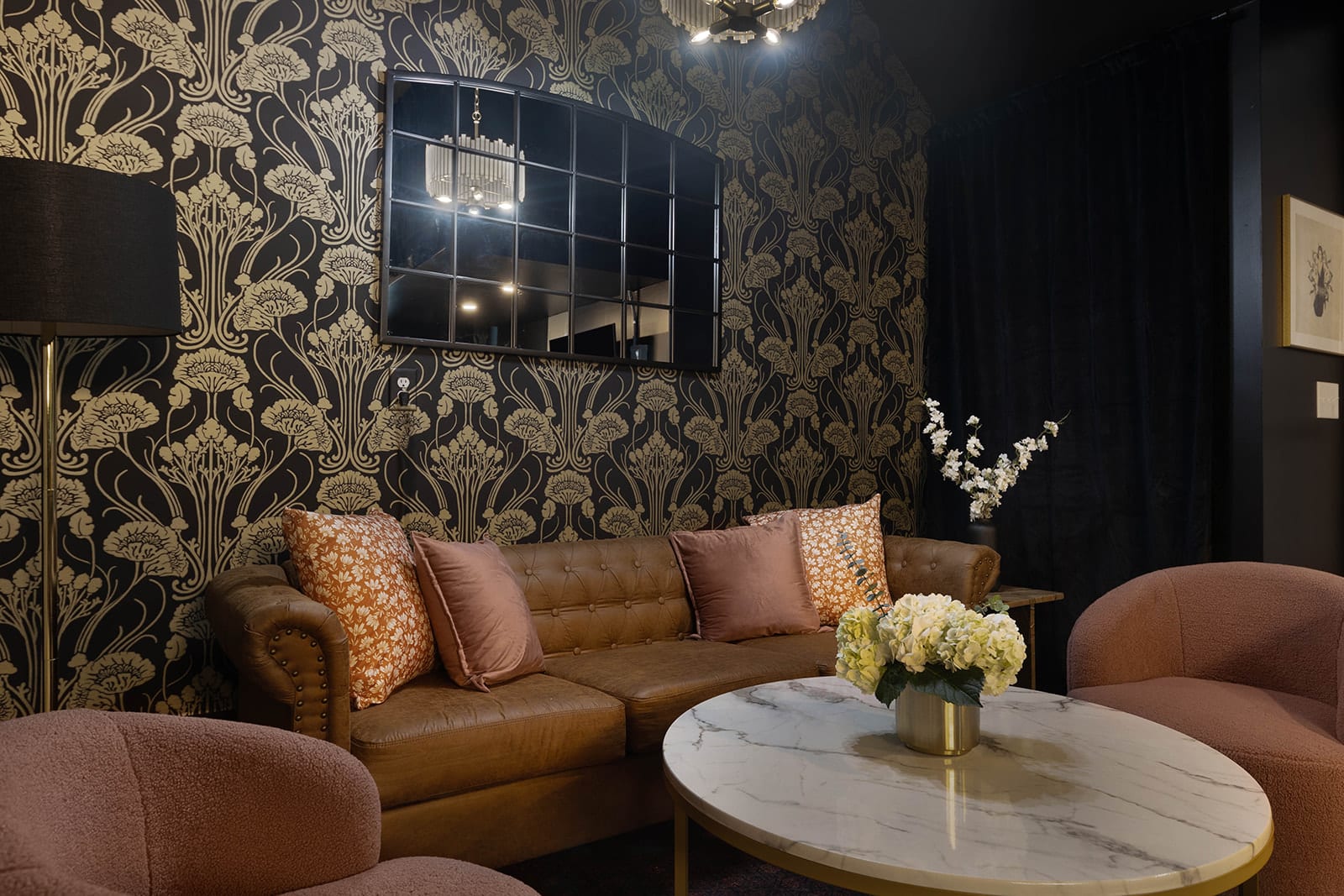 A close-up of the elegant living room showcases a brown tufted leather sofa adorned with pink and patterned pillows, a round marble coffee table with a floral centerpiece, and two plush armchairs.