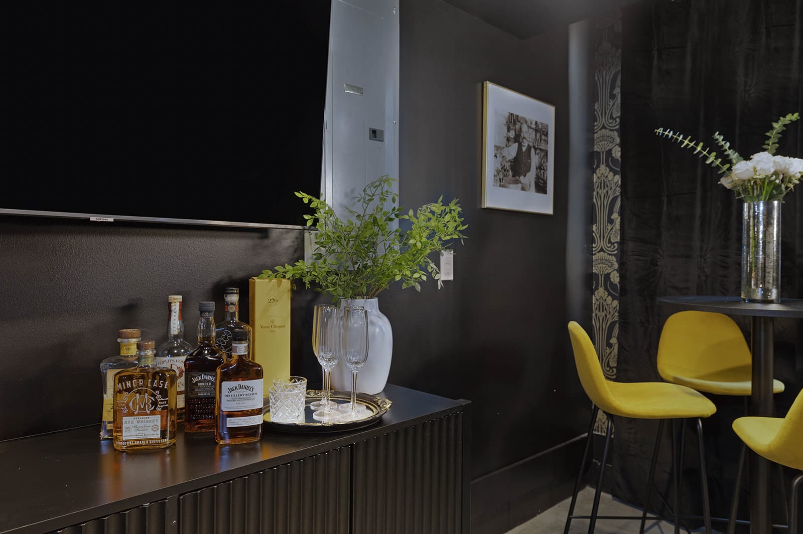A detailed view of the speakeasy's bar area features a dark wood cabinet with various bourbon bottles, champagne flutes, a silver tray, and a vase with greenery, all beneath a wall-mounted TV.