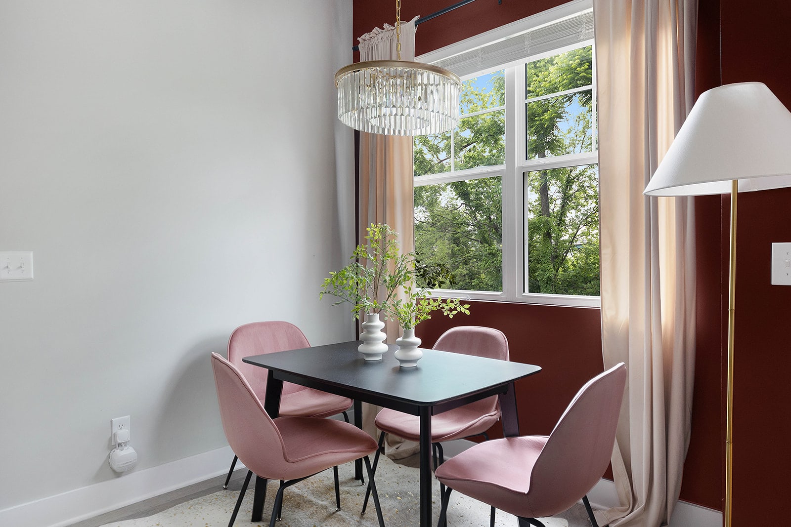 A cozy dining nook features a black rectangular table and four pink velvet chairs, set against a white wall with a red accent section. A large window offers views of lush greenery, and a modern crystal chandelier hangs above the table.