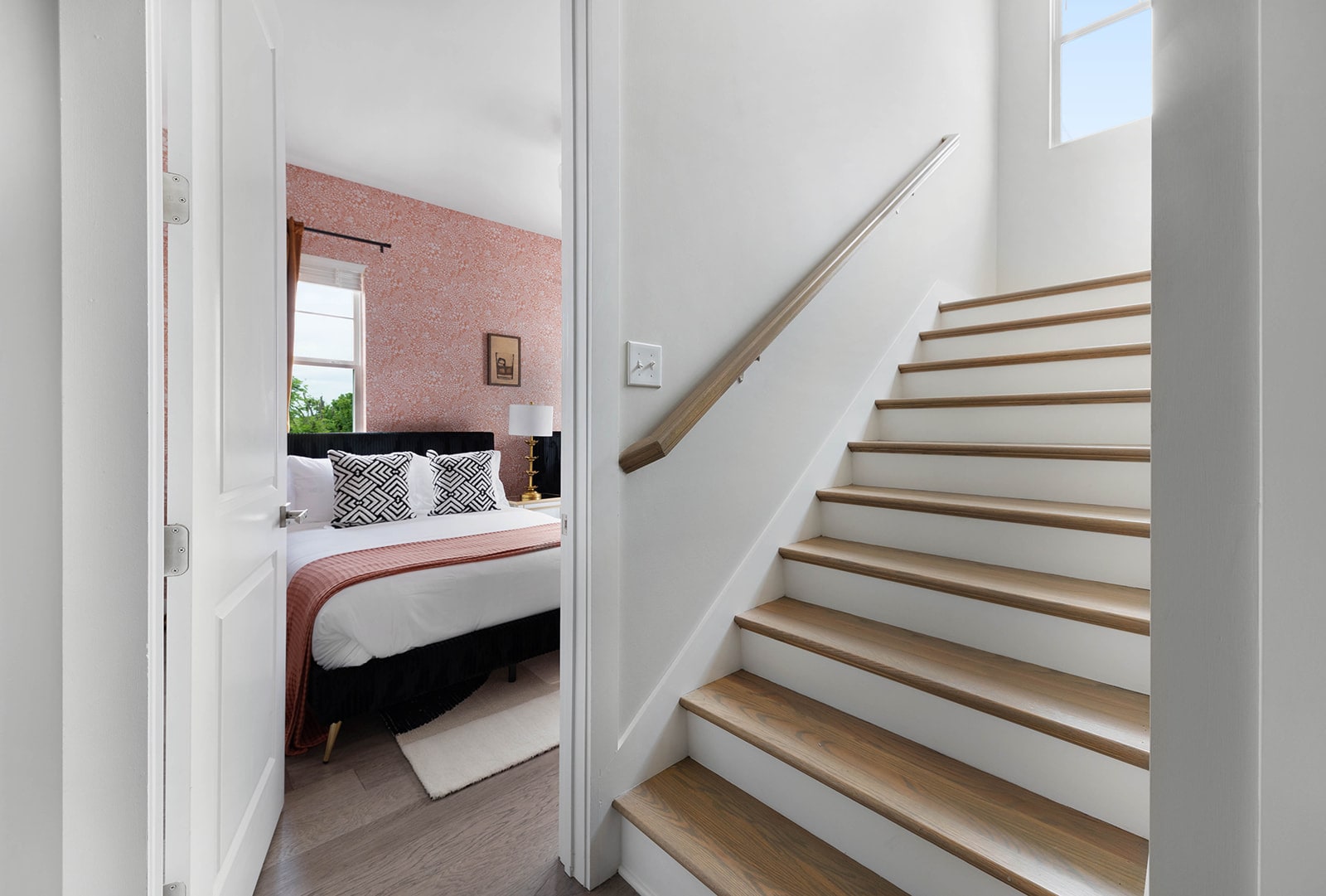 A bright interior staircase with light wood treads and white risers, leading up to a landing. A bedroom with a patterned accent wall and a bed with a black headboard and pink bedding is visible through an open doorway.