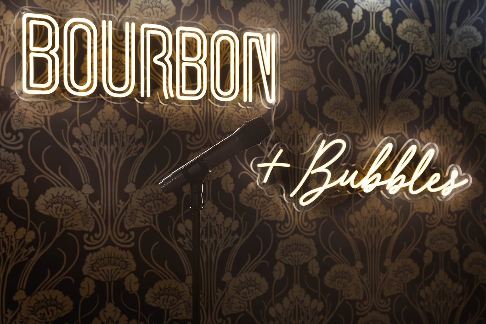 A detailed close-up of the "BOURBON + Bubbles" neon signs illuminating the ornate black and gold wallpaper in the speakeasy-themed entertainment room, with a microphone stand in the foreground.