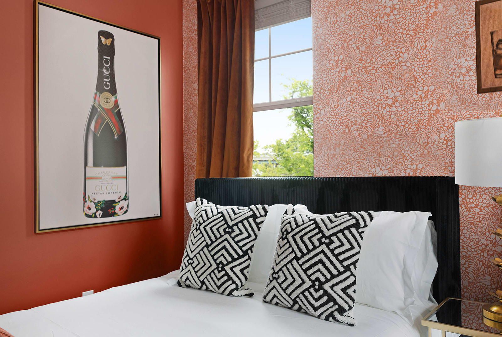 A close-up of one bed in a stylish bedroom, featuring black and white patterned pillows and a striking large framed artwork of a 'Gucci' champagne bottle on the wall.