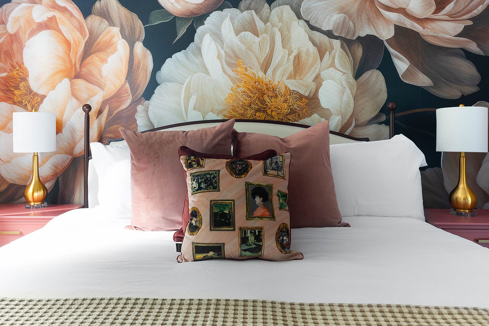 A close-up of a bed in a bedroom adorned with a dramatic floral mural, featuring plush pink velvet pillows and a unique decorative throw pillow with framed portraits. Gold lamps sit on elegant pink nightstands.