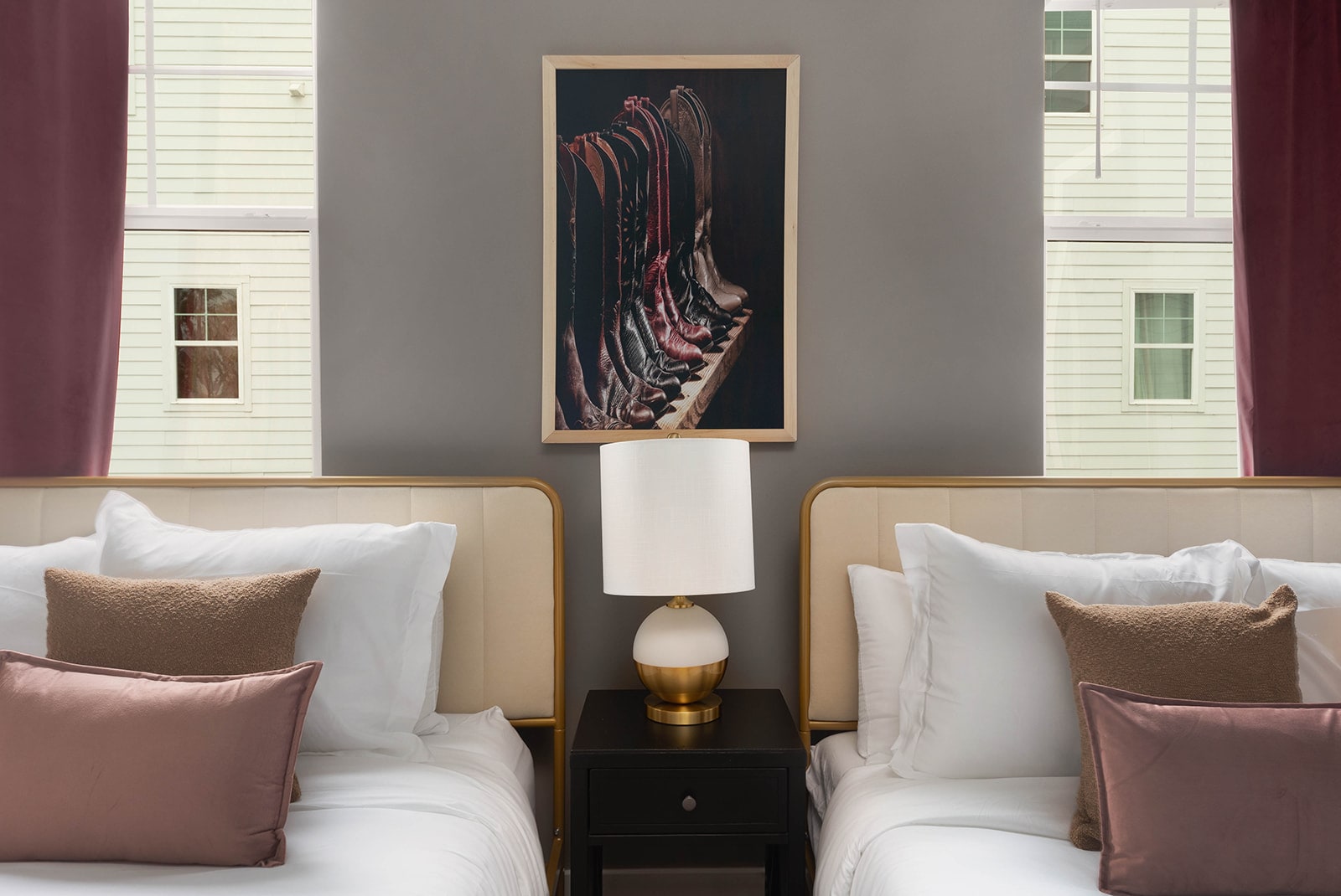 A bedroom features two twin beds with cream upholstered headboards and gold frames, separated by a dark nightstand with a white and gold lamp. A large framed art print of cowboy boots hangs on the gray wall, with burgundy curtains framing the windows.