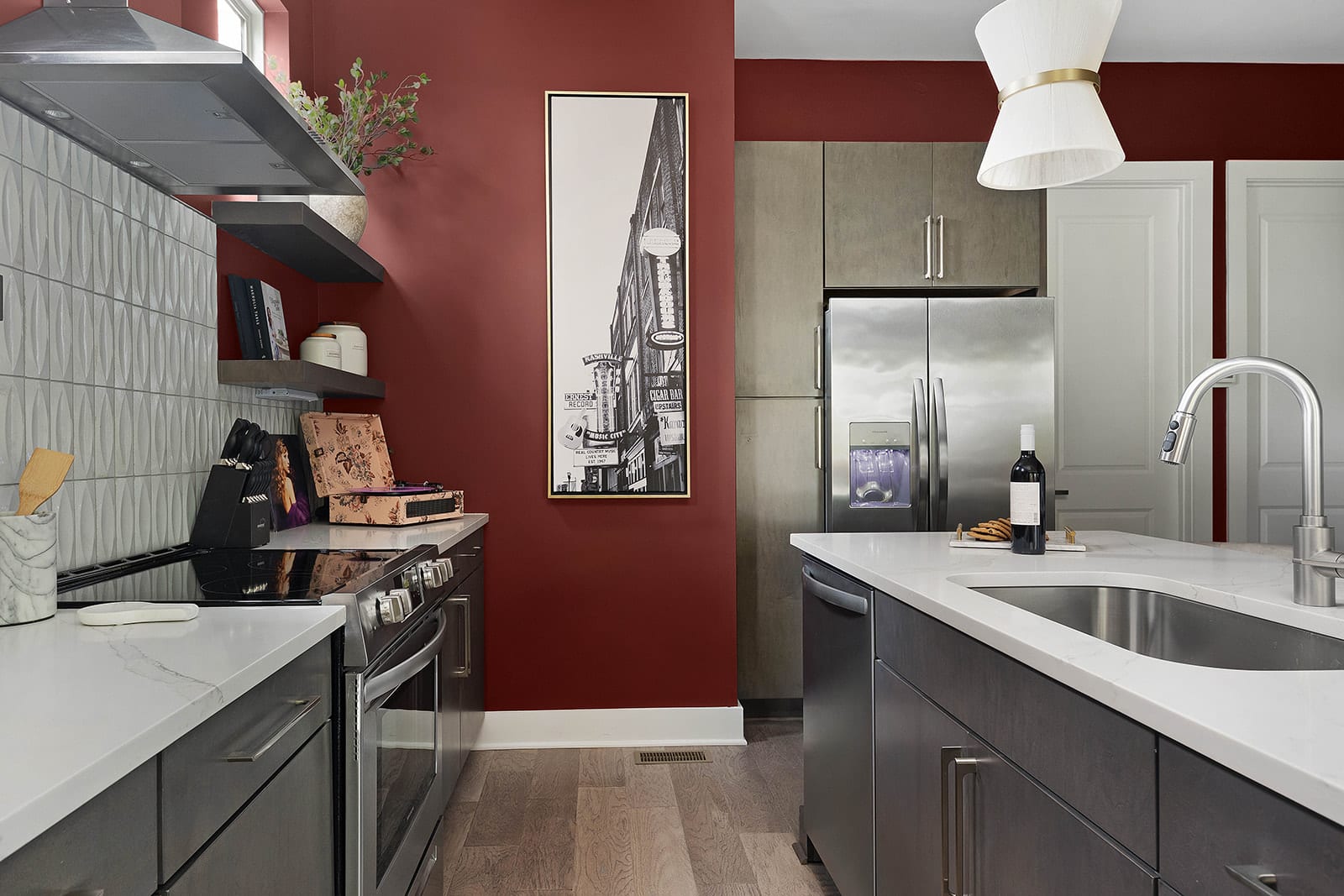 A modern kitchen showcases dark gray cabinetry, white quartz countertops, and a striking red accent wall. Stainless steel appliances, including a refrigerator and range, are visible alongside a tall black and white framed art piece.