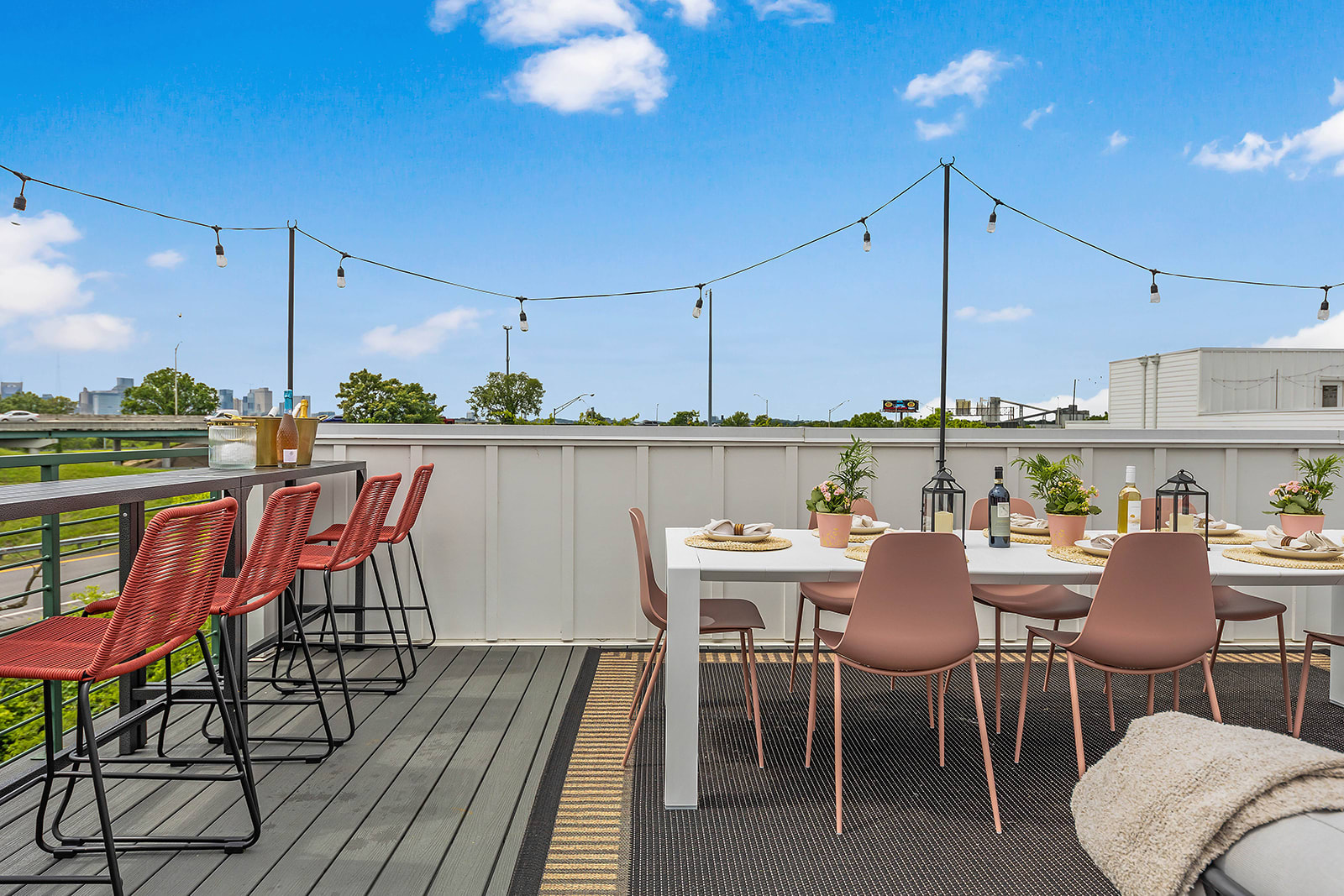 A sunny rooftop deck with an outdoor dining area featuring a long white table with pink chairs, string lights, and a bar area with red stools. The deck overlooks distant cityscapes under a clear blue sky.