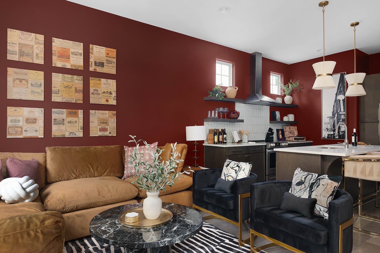 An expansive open-concept area showcases a modern kitchen with red walls, dark cabinetry, a large island, and a living room with a brown sectional sofa and dark blue armchairs.