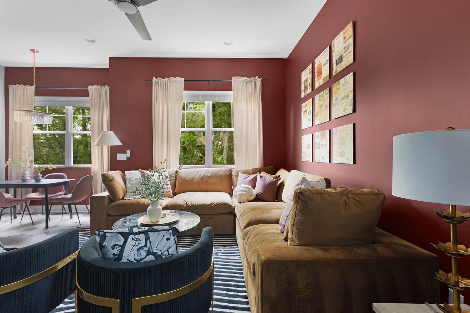 An open-concept living space with red walls features a large brown sectional sofa, two dark blue armchairs, a round coffee table, and a dining area with a round table and pink chairs visible in the background.