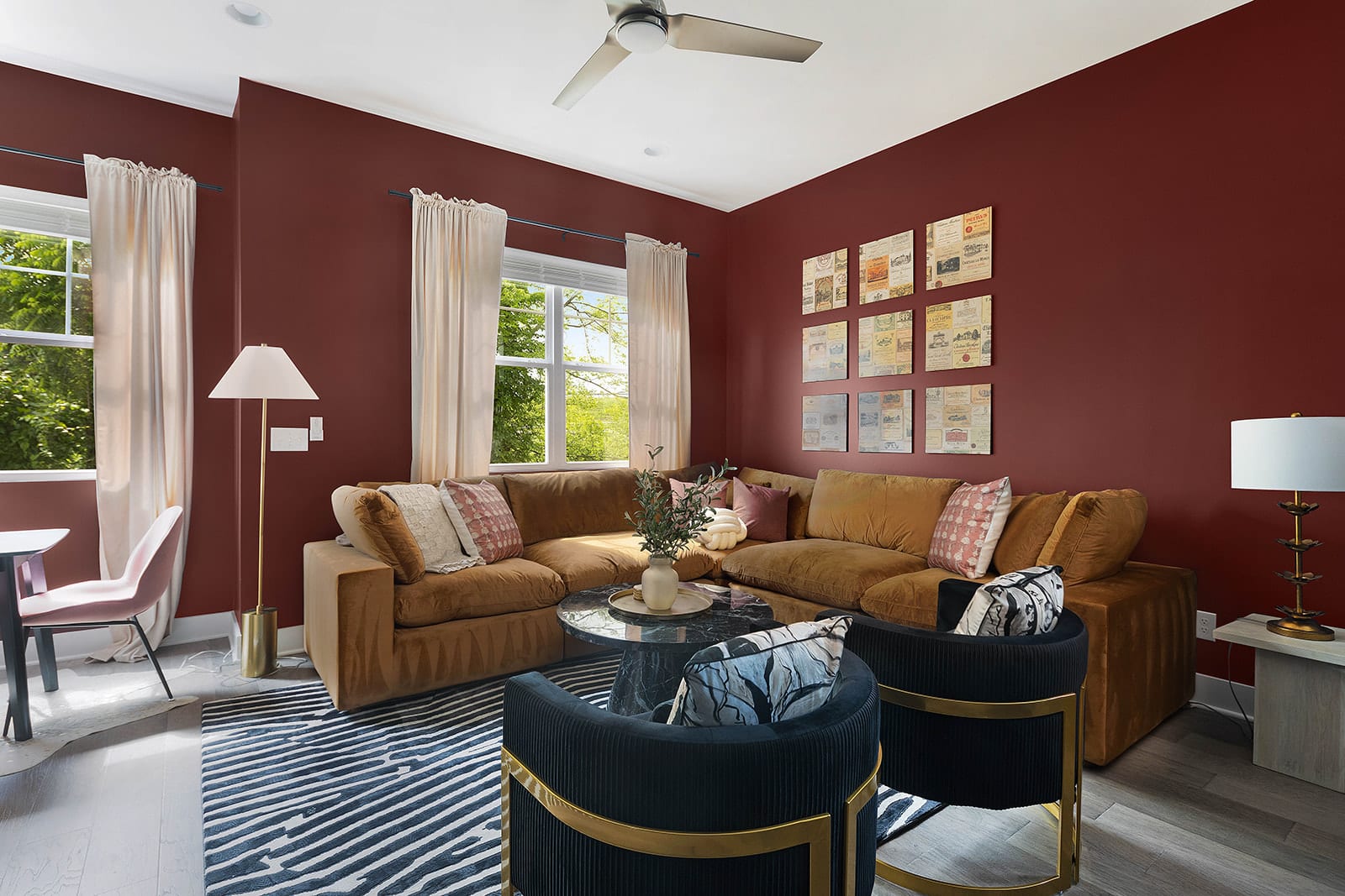 A vibrant living room with rich red walls features a large L-shaped brown sectional sofa, two dark blue armchairs with gold accents, a round black marble coffee table, and a striped rug.