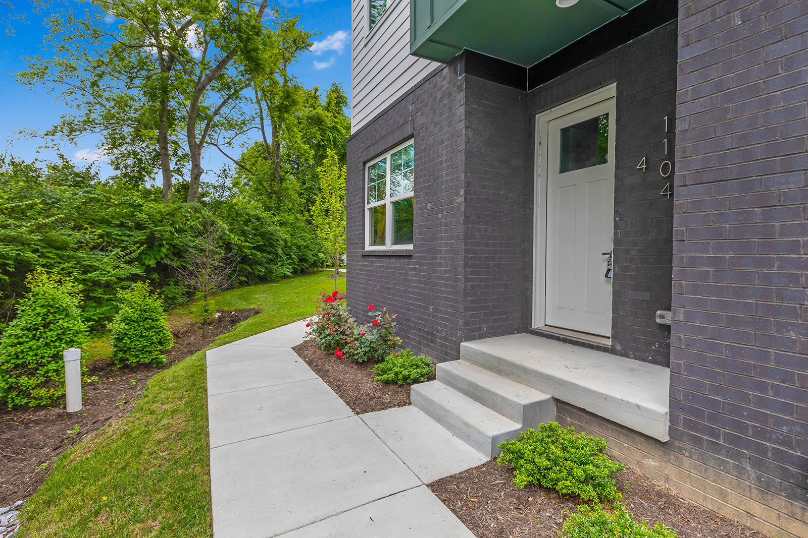 Front entrance of a modern townhouse with dark brick and green siding, a white door, concrete steps, and landscaped garden beds with green shrubs and red flowers.
