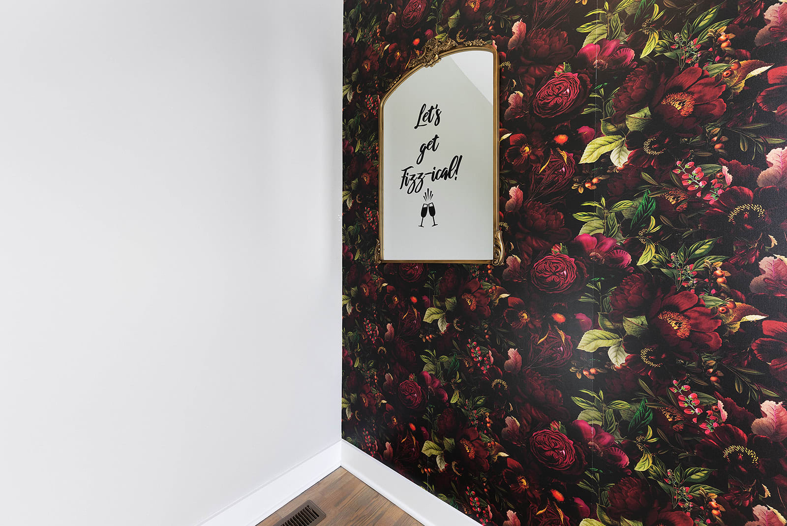 A vibrant wall covered in dark floral wallpaper with deep red roses and green foliage, featuring a gold-framed arched mirror reflecting a "Let's get Fizz-ical!" sign. A white wall is visible on the left, and a wood floor vent is at the bottom.