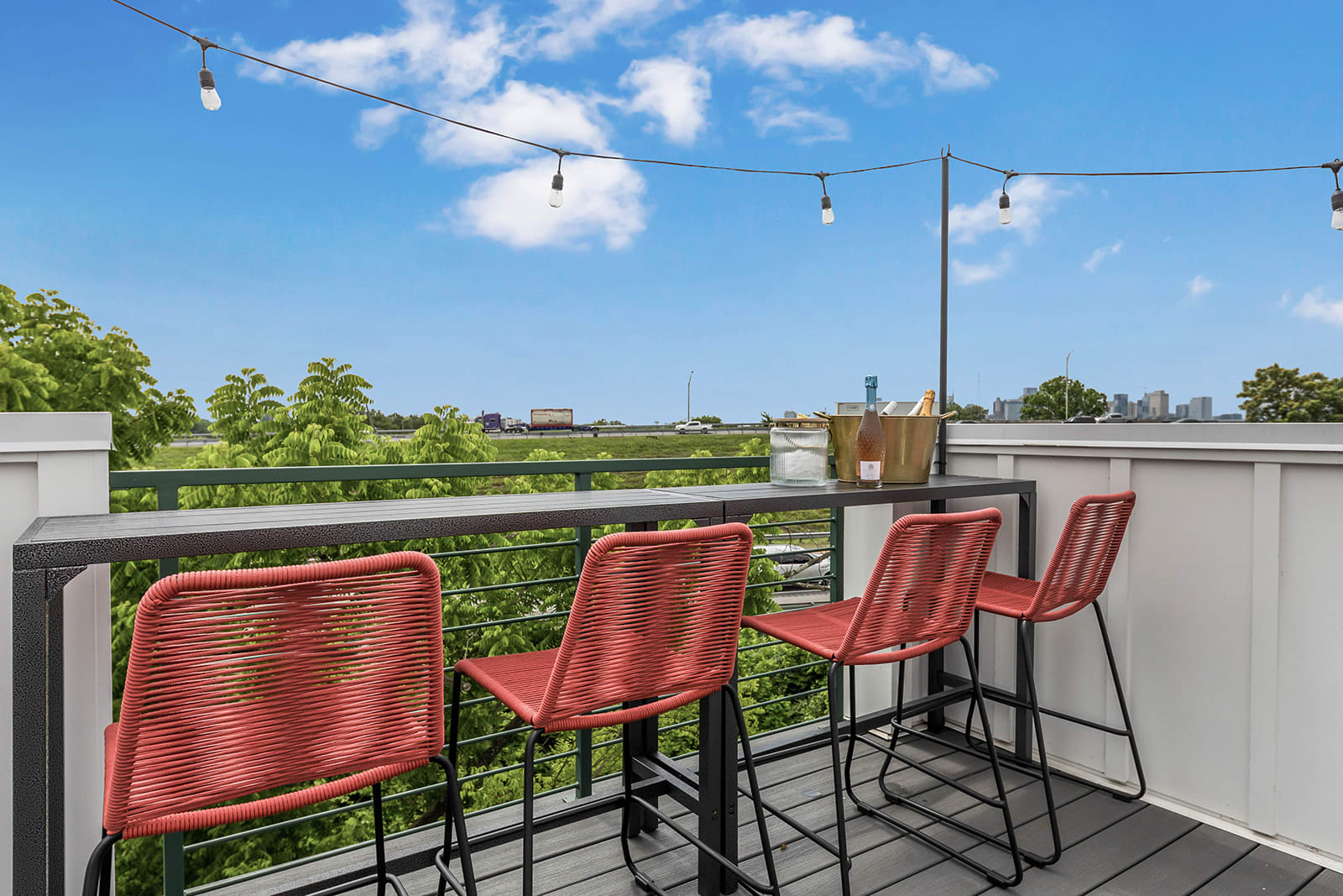 Rooftop deck with a long bar table and three red woven bar stools, string lights, and a clear blue sky with a distant city view and highway visible through lush green trees.