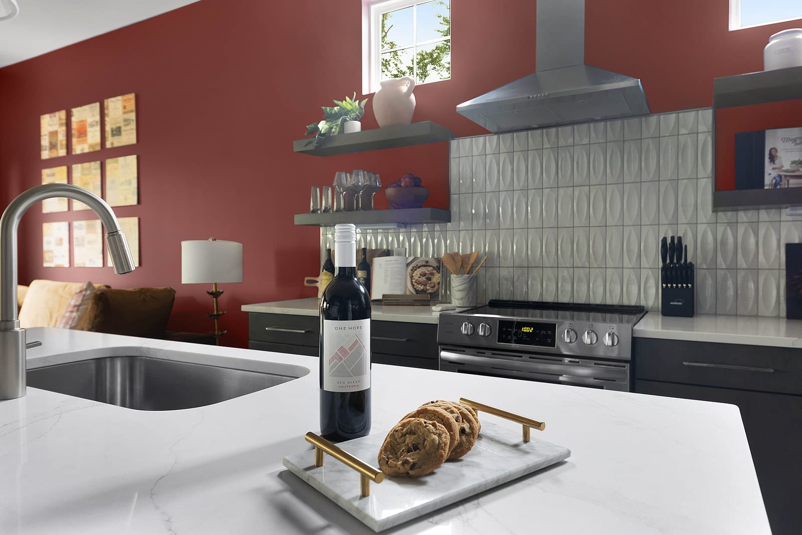 A bright kitchen features a bold red accent wall, white quartz countertops, and a stainless steel range with a matching hood. A bottle of red wine and a tray of cookies are invitingly placed on the island, complemented by open shelving with glassware.